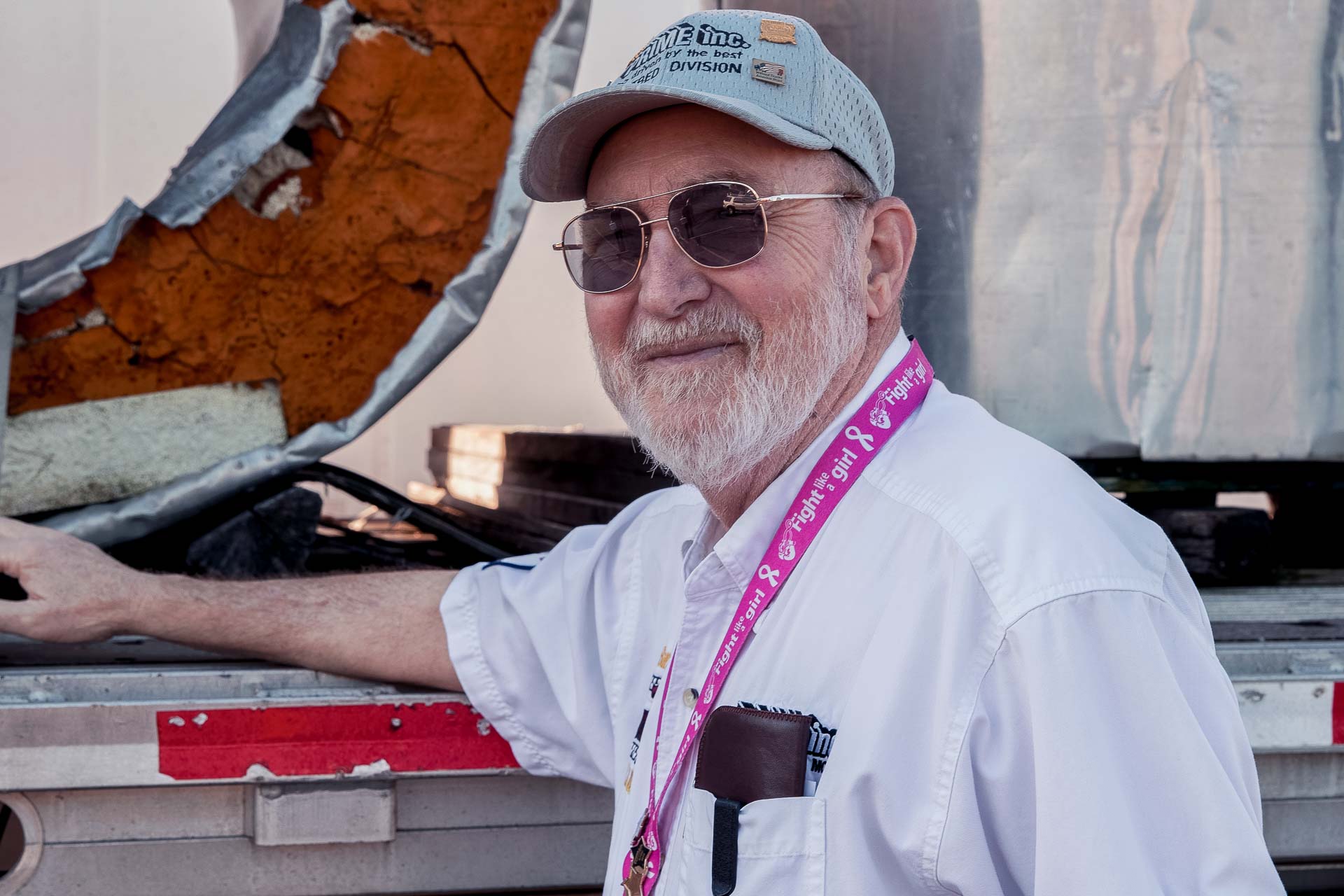 A Prime truck driver, wearing a grey hat and sunglasses, standing next to a flatbed semi-trailer.